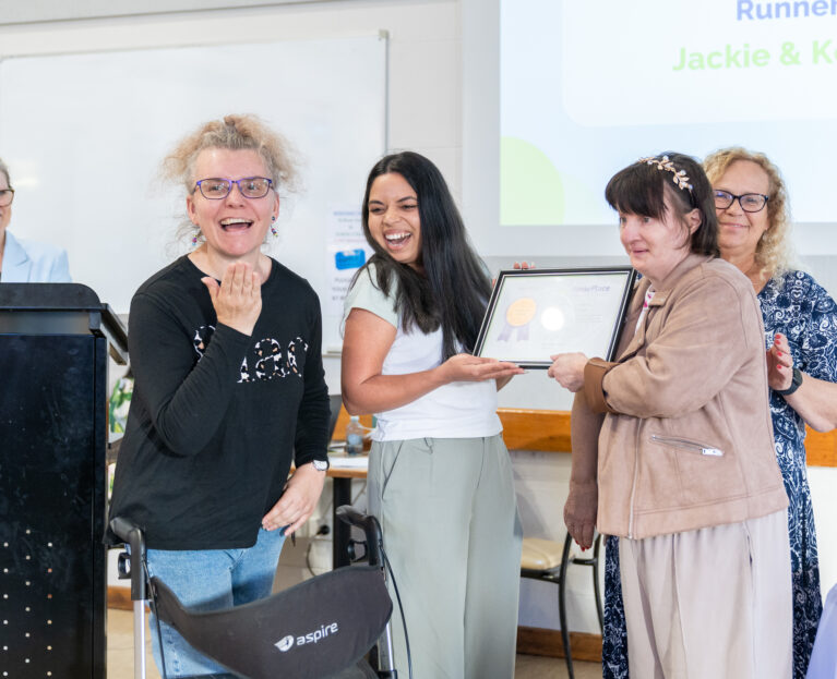 A HomePlace worker being recognised for her hard work. She is smiling and holding her certificate with HomePlace participants.