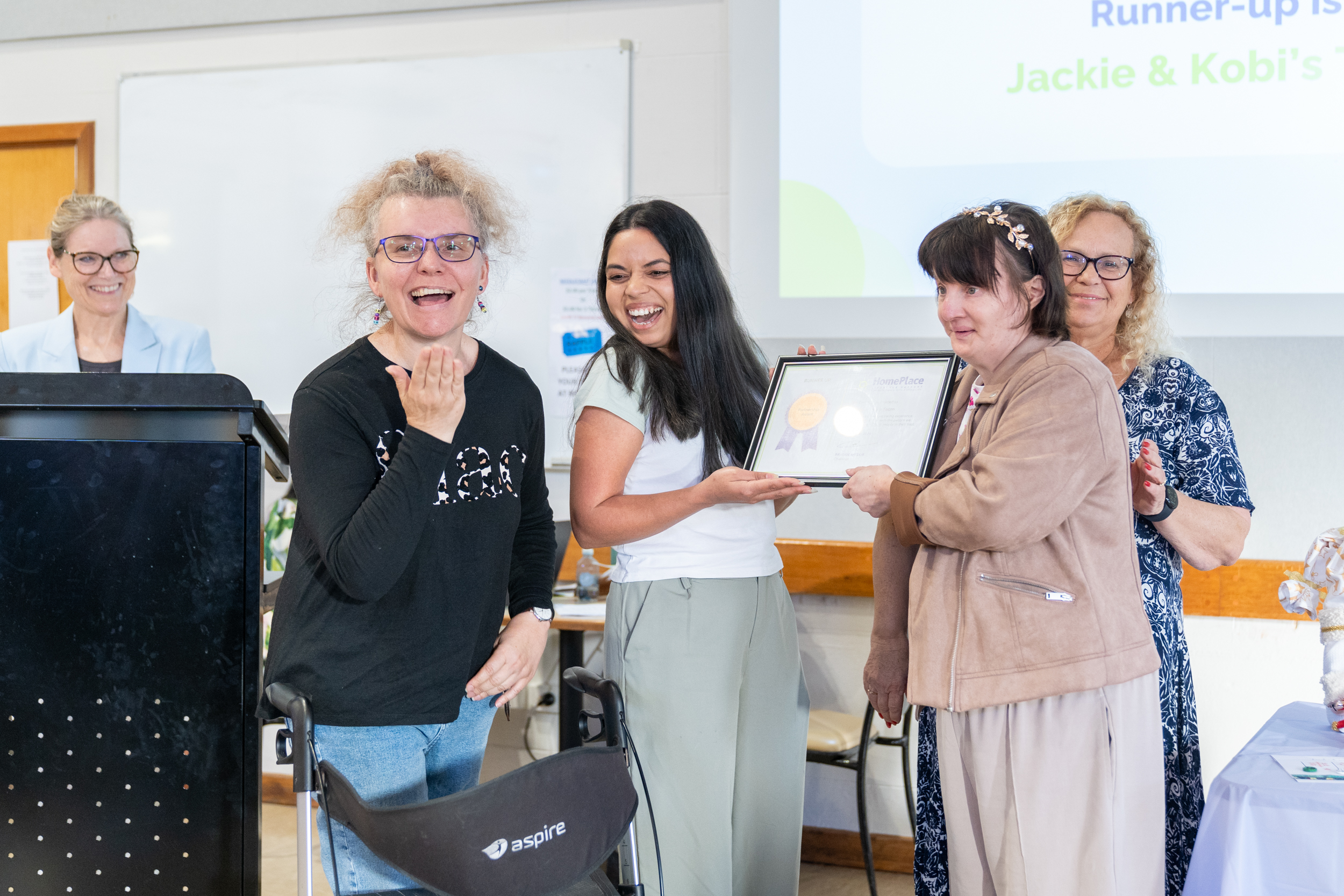 A HomePlace worker being recognised for her hard work. She is smiling and holding her certificate with HomePlace participants.