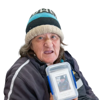 Participant Debbie wearing a blue and black knitted hat and a black jumper. She is smiling and holding up a container with some cards.
